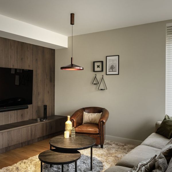 Modern living room with a wall-mounted TV, leather armchair, round wooden tables, beige sofa, and minimal decor on neutral walls.