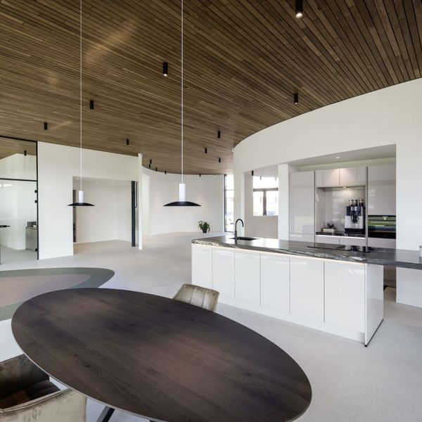 Modern open-plan kitchen and dining area with curved wood ceiling, sleek white cabinets, round table, and large windows for natural light.