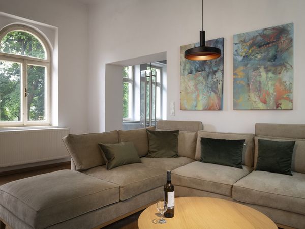 Cozy living room with a beige sectional sofa, abstract artwork on the wall, a round wooden table with a wine bottle and glass, and a large arched window.