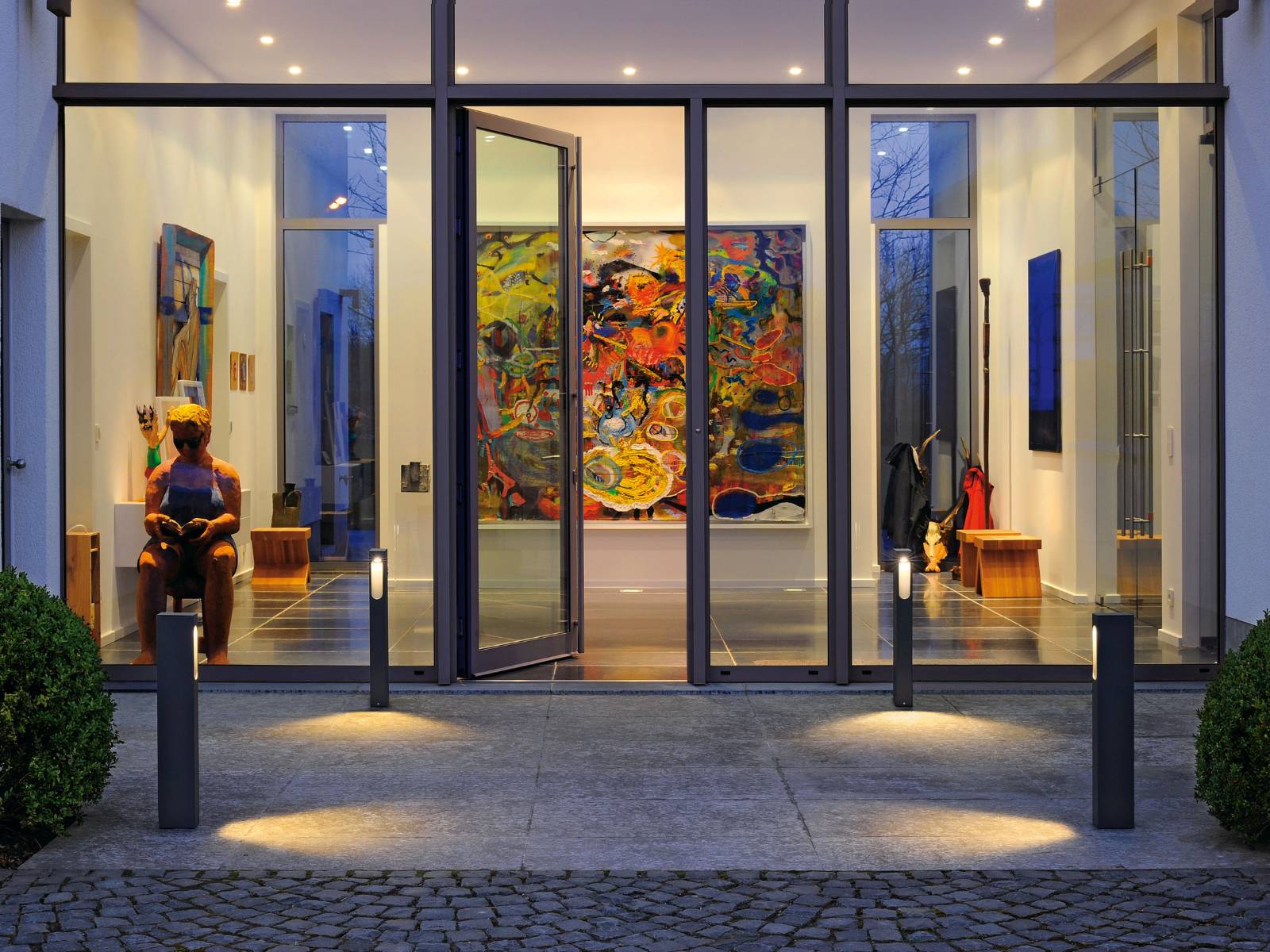 Beleuchteter Glas-Eingangsbereich mit Pollerleuchten auf dem Vorplatz, der den Blick auf farbenfrohe Kunstwerke im Inneren freigibt