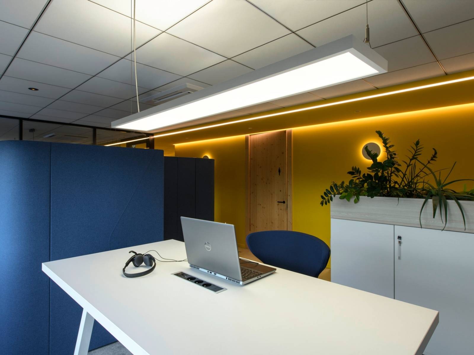 Moderner Büro-Arbeitsplatz mit einem Dell-Laptop auf einem weißen Schreibtisch, einer blauen Trennwand, gelber Akzentwand mit LED-Beleuchtung und einer großen linearen Pendelleuchte.