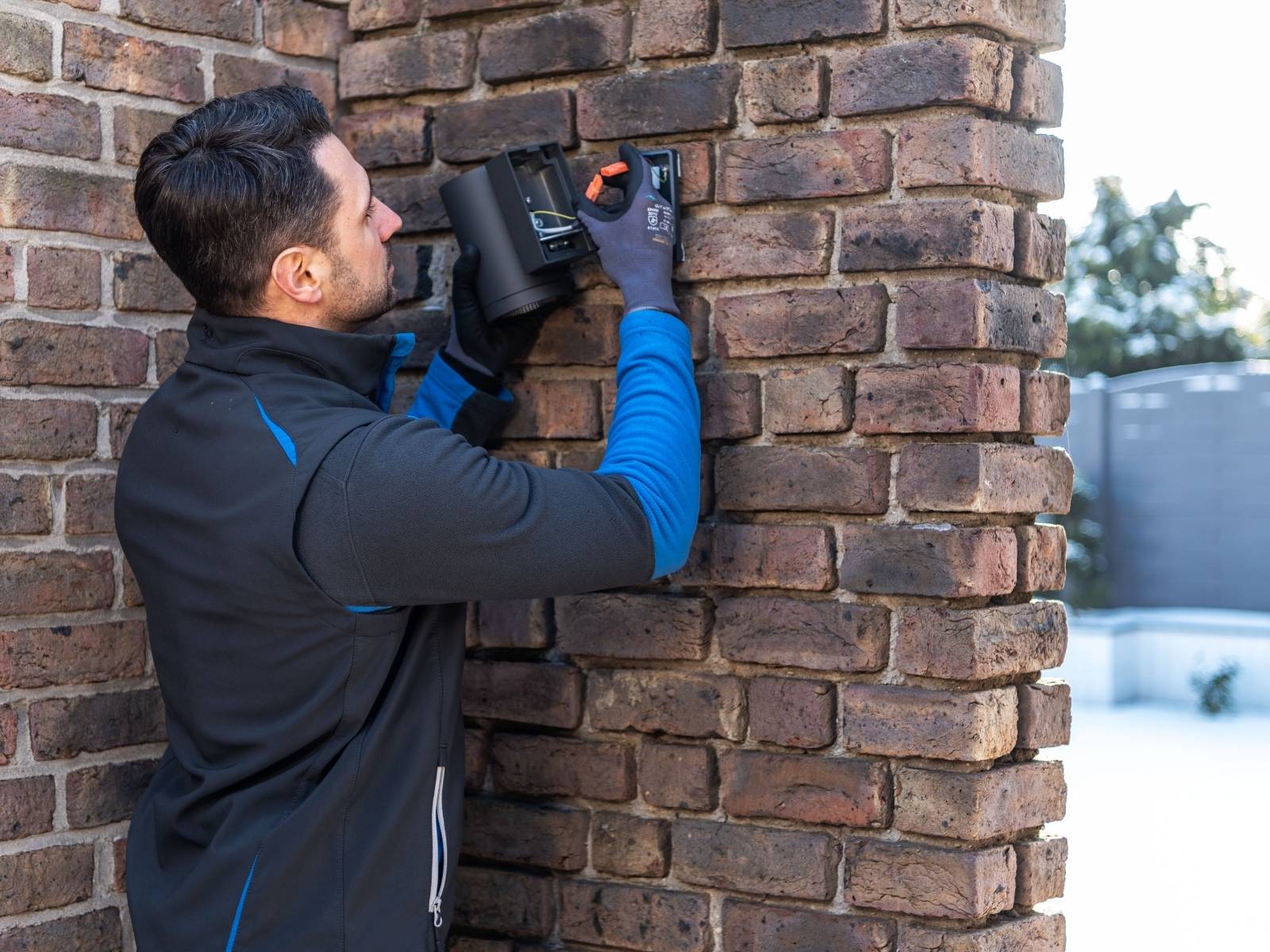 Elektriker, der eine Wandleuchte an einer Backsteinmauer anbringt und eine schwarz-blaue Jacke sowie Handschuhe trägt. Außenaufnahme mit Bäumen im Hintergrund.