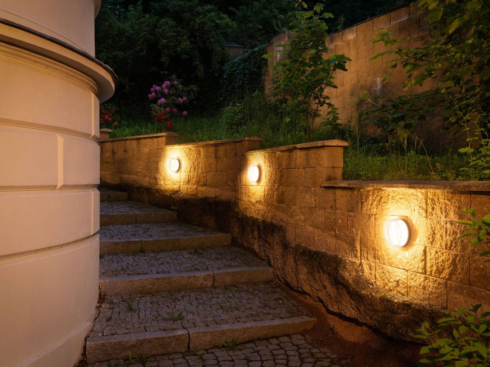 Außenliegende Steintreppe bei Nacht, die an einer Natursteinmauer hinaufführt, welche durch runde, warmweiße Wandeinbauleuchten sicher erhellt wird.