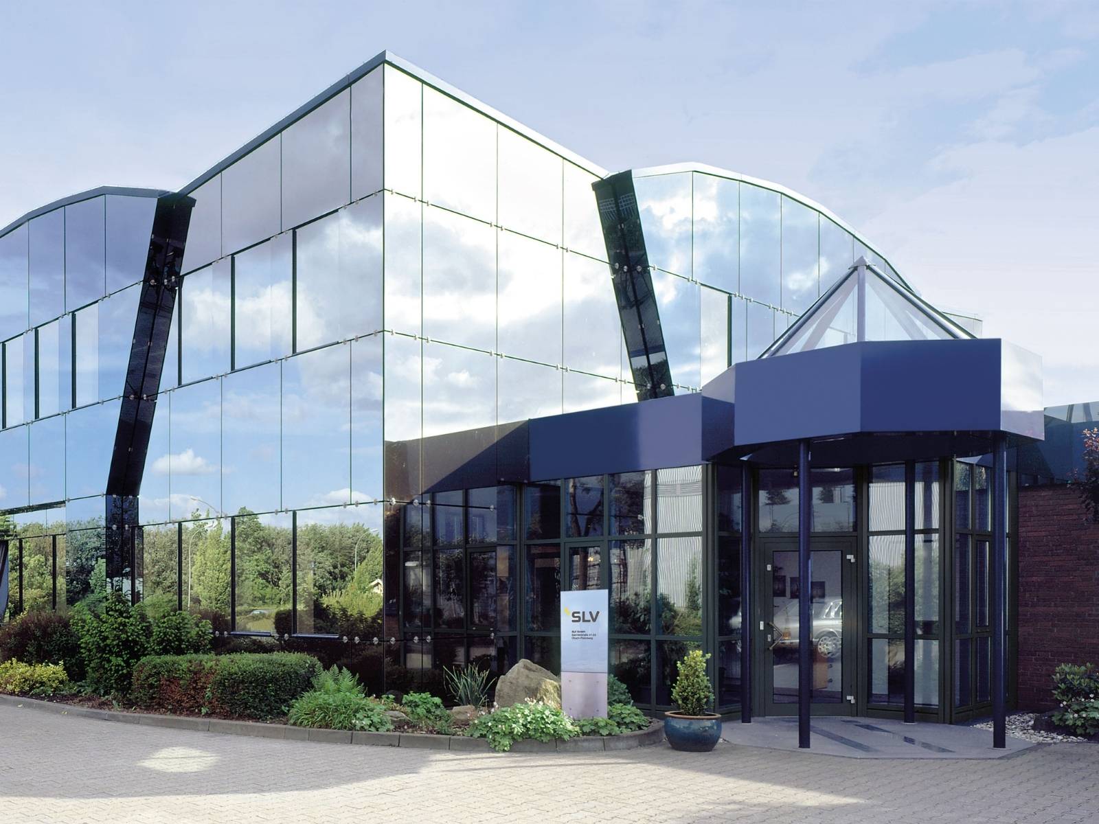Der moderne Hauptsitz der SLV GmbH in Übach-Palenberg mit einer großen, spiegelnden Glasfassade, einem blauen Eingangsbereich und einer SLV-Firmensäule im Vordergrund.