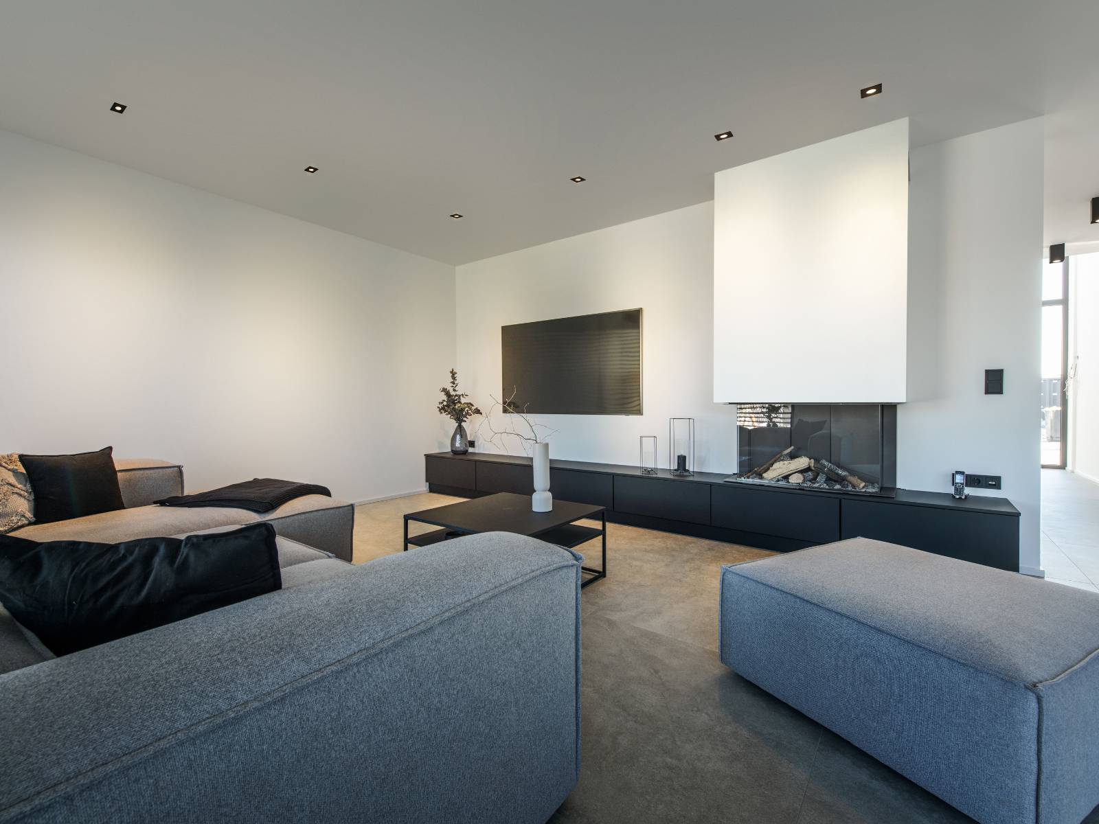 Dezente quadratische schwarze Einbauleuchten in einer weißen Wohnzimmerdecke über einer modernen Einrichtung mit großem grauem Sofa und Kamin