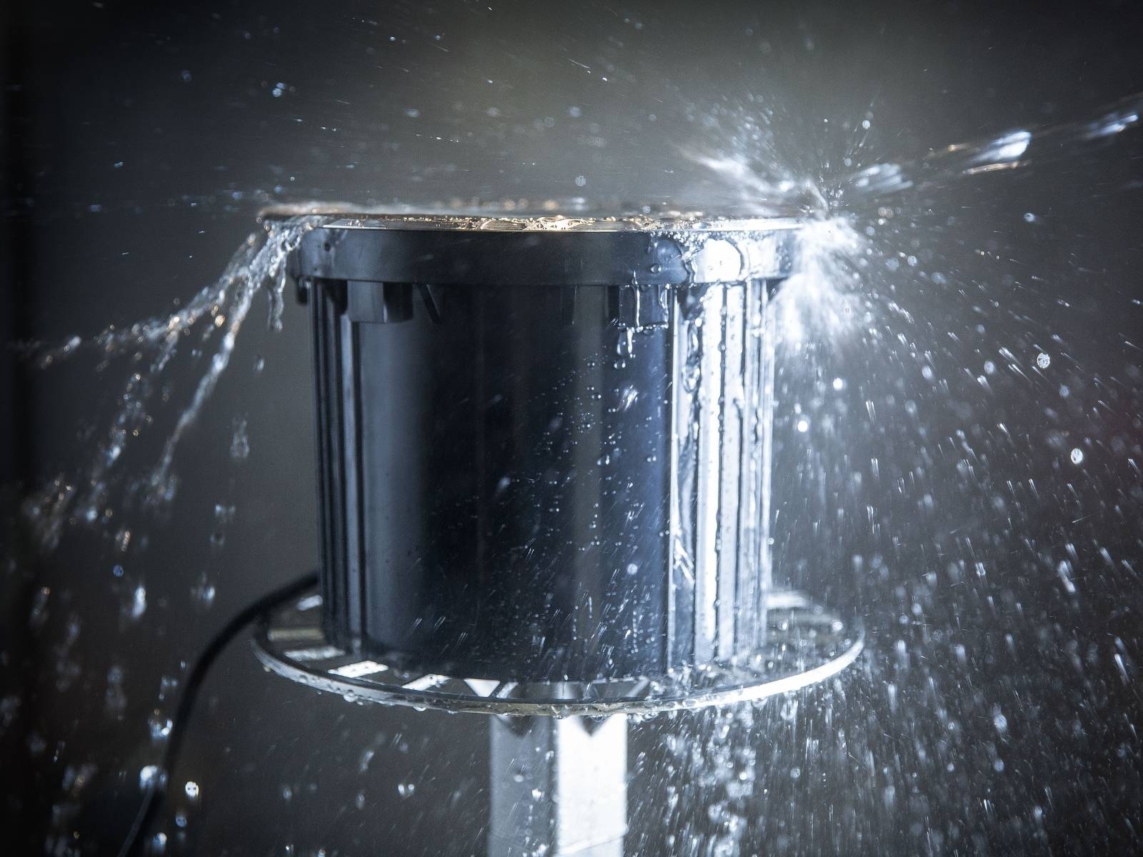 Wasser spritzt auf eine Einbauleuchte in einer Testkammer, vor einem dunklen Hintergrund.