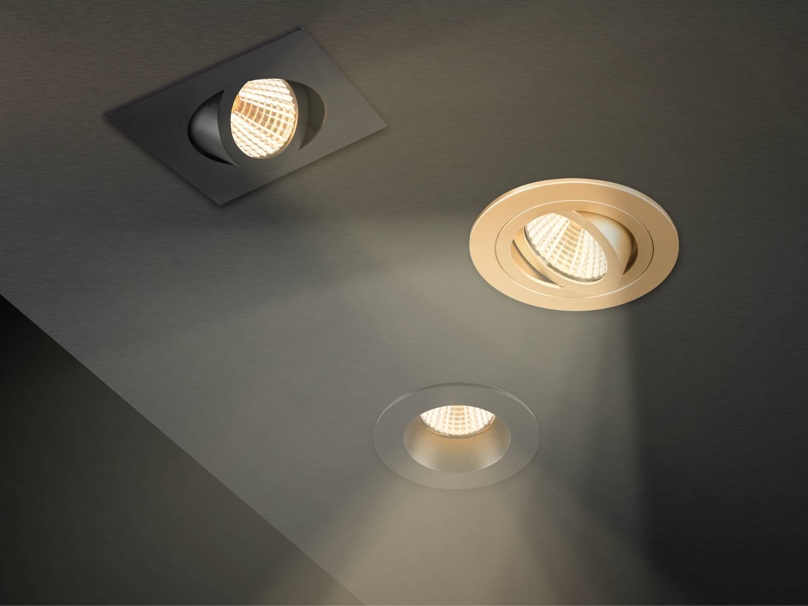 Drei verschiedene quadratische und runde LED-Einbaustrahler in Schwarz, Gold und Silber an einer grauen Decke mit warmweißem Lichtkegel.