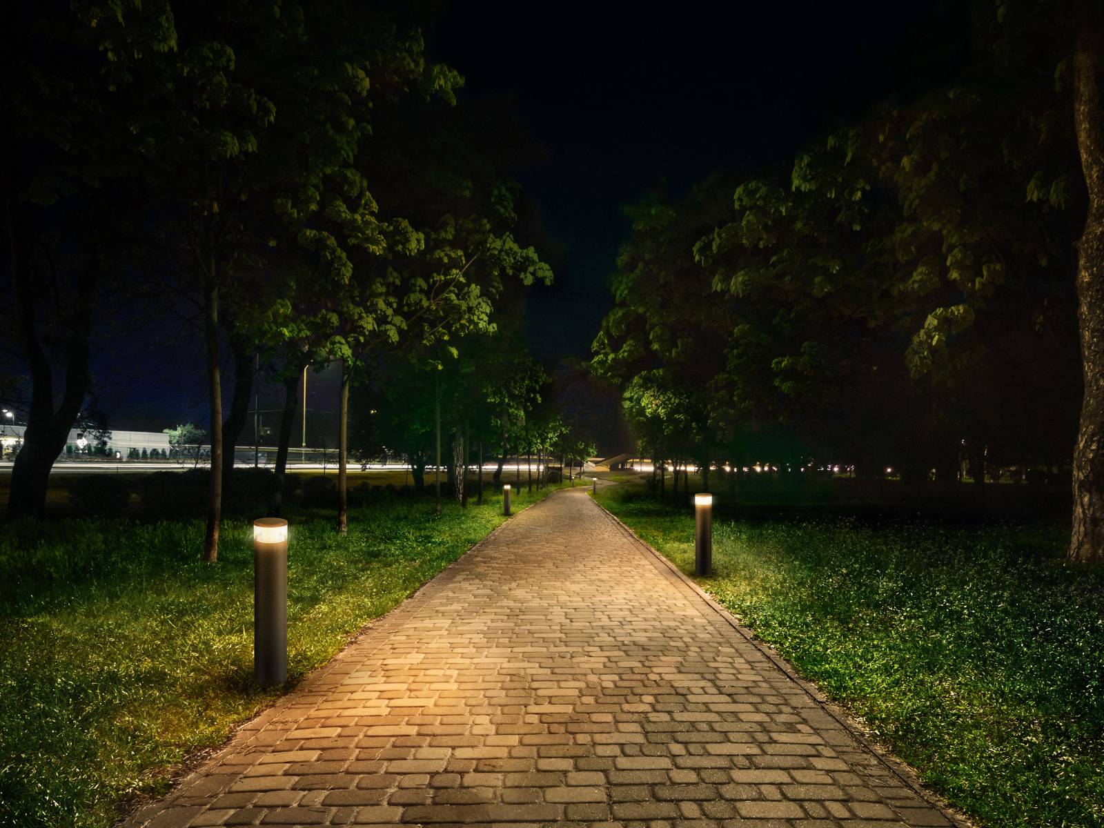 Ein beleuchteter Kopfsteinpflasterweg durch einen Park bei Nacht, gesäumt von Bäumen und M-POL Standleuchten, der in die Ferne führt.