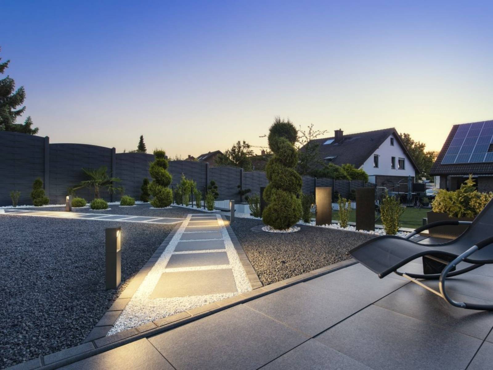 Modern gestalteter Garten mit anthrazitfarbenem Kies, Steinplattenweg und eckigen LED-Pollerleuchten, die den Weg am Abend hell ausleuchten.