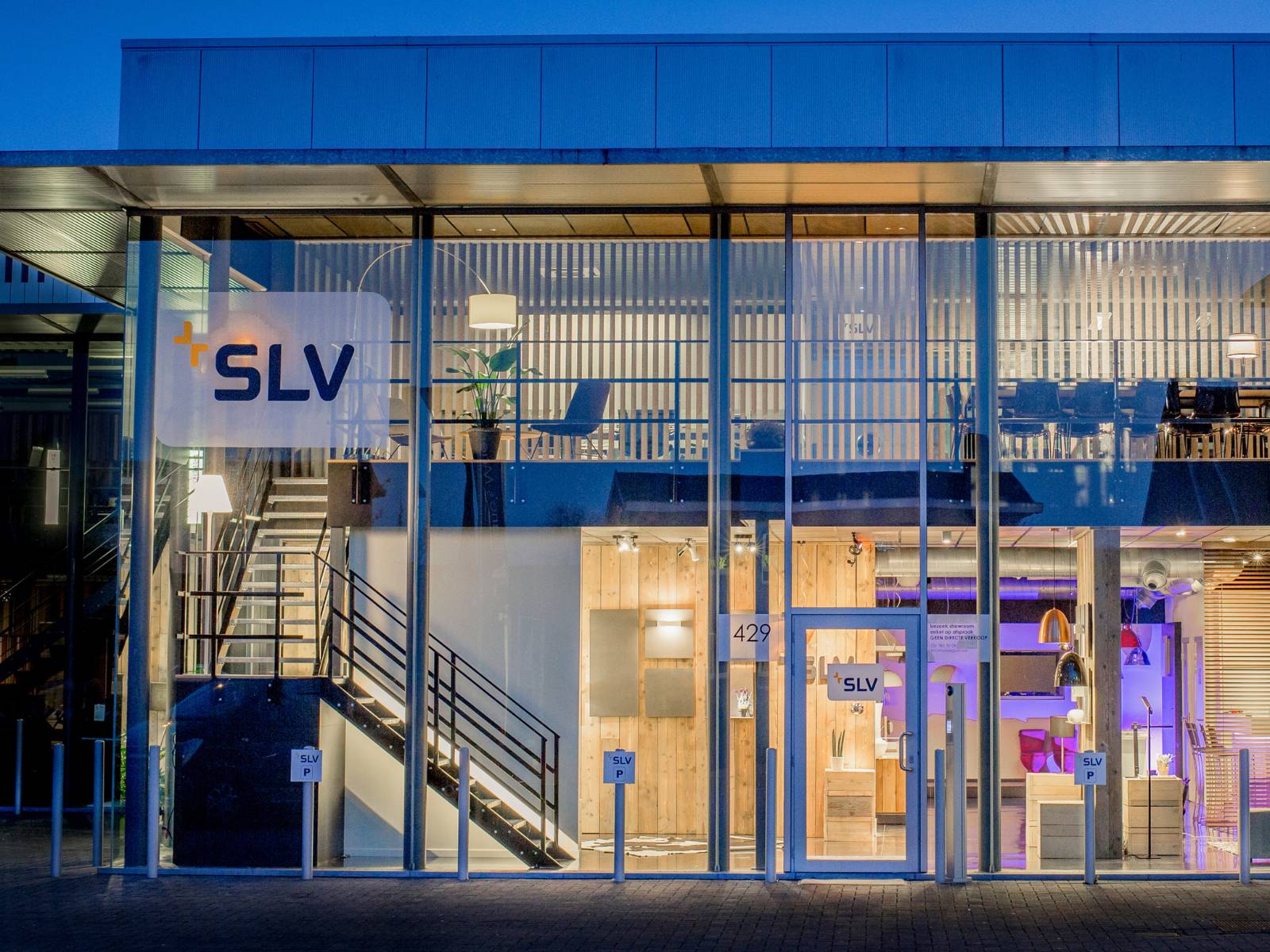 Bâtiment de la société SLV Belgique : vue extérieure du bâtiment SLV au design moderne à la tombée de la nuit, avec une façade en verre transparente qui permet d'apercevoir le showroom éclairé et l'étage des bureaux