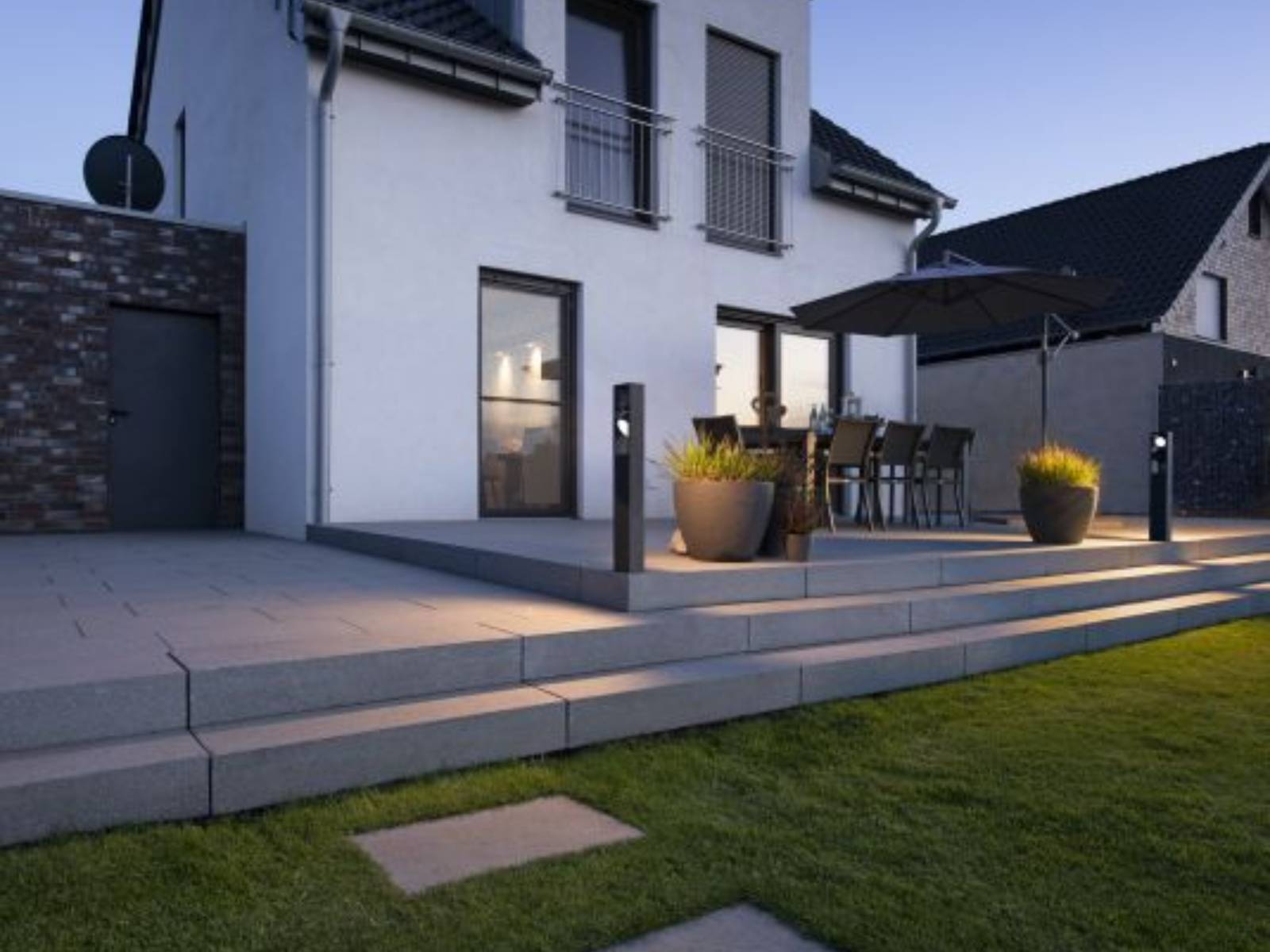 Moderne Terrasse mit Stehleuchten, eleganten Sitzgelegenheiten im Freien, großen Topfpflanzen und einem Sonnenschirm auf stilvoll beleuchteten Stufen. Im Hintergrund ein modernes zweistöckiges Haus.