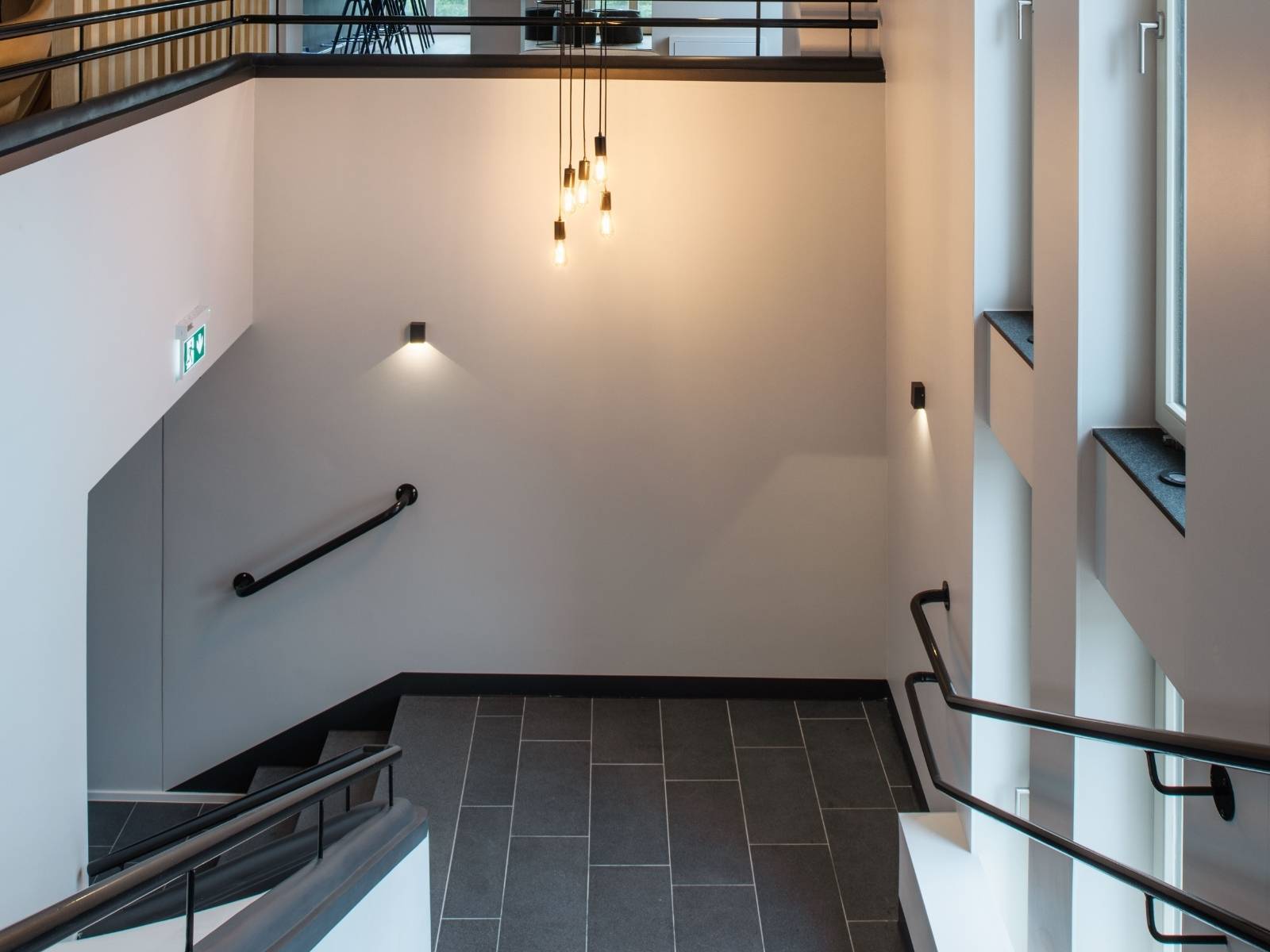 Modernes Treppenhaus mit dunklen Fliesen, weißen Wänden, schwarzen Geländern und einer mehrflammigen, von der Decke herabhängenden Pendelleuchte mit dekorativen Glühbirnen.