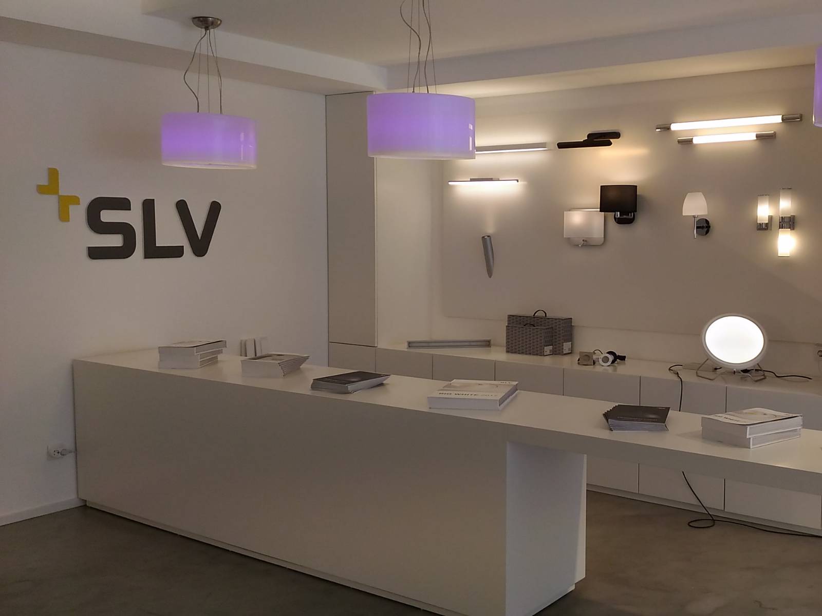 Showroom moderno e iluminado com balcão de atendimento branco, catálogo BIG WHITE em exposição, logótipo da SLV na parede e várias luminárias de teto e de parede acesas.
