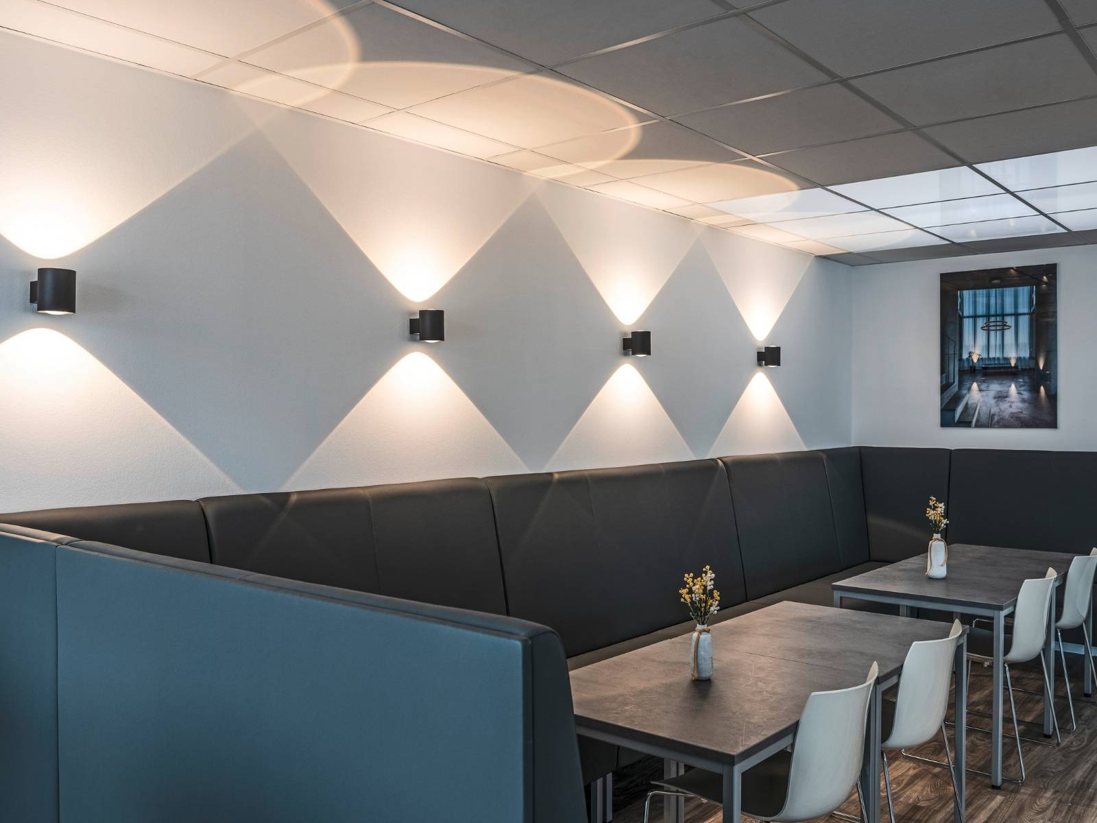 Vier schwarze, quadratische Up-and-Down-Wandleuchten erzeugen dekorative, sanduhrförmige Lichtkegel an einer weißen Wand in einer Sitzecke.