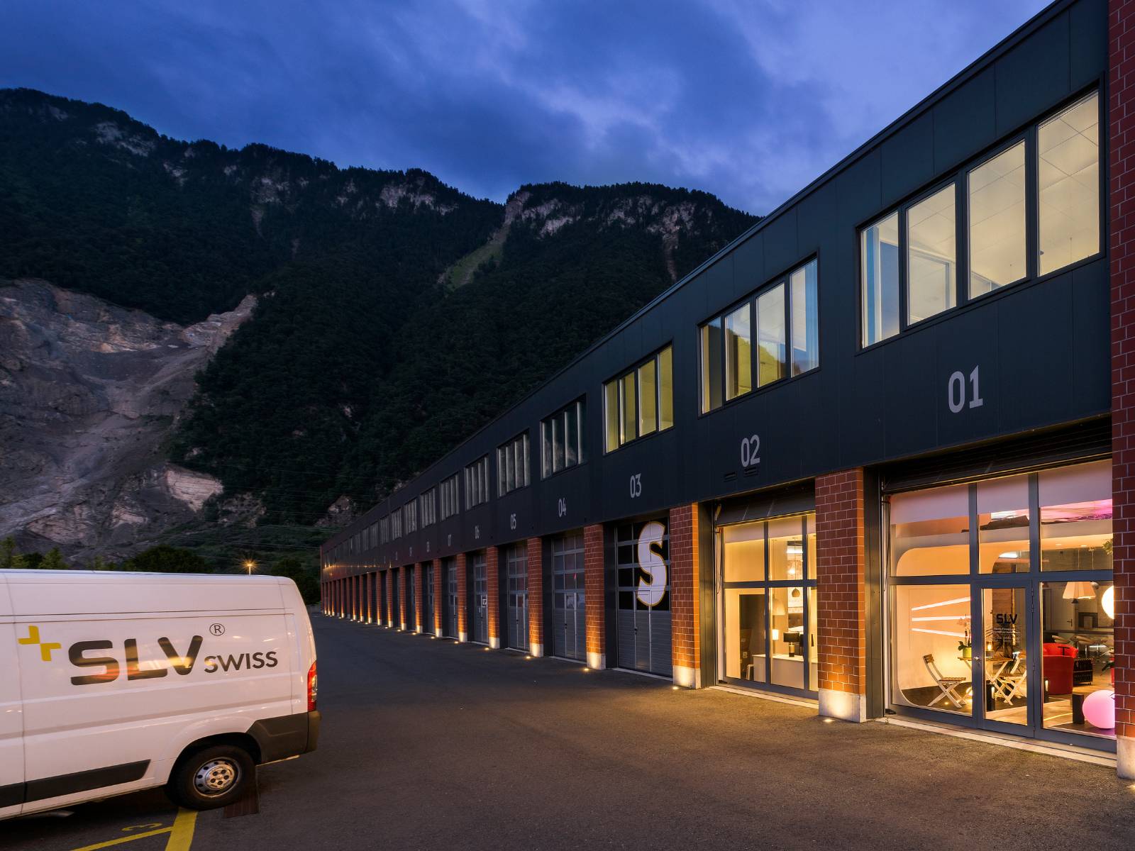Außenansicht des SLV Swiss Gebäudes bei Dämmerung mit beleuchteten Fensterfronten, einem weißen Service-Fahrzeug im Vordergrund und einer eindrucksvollen Gebirgskulisse im Hintergrund