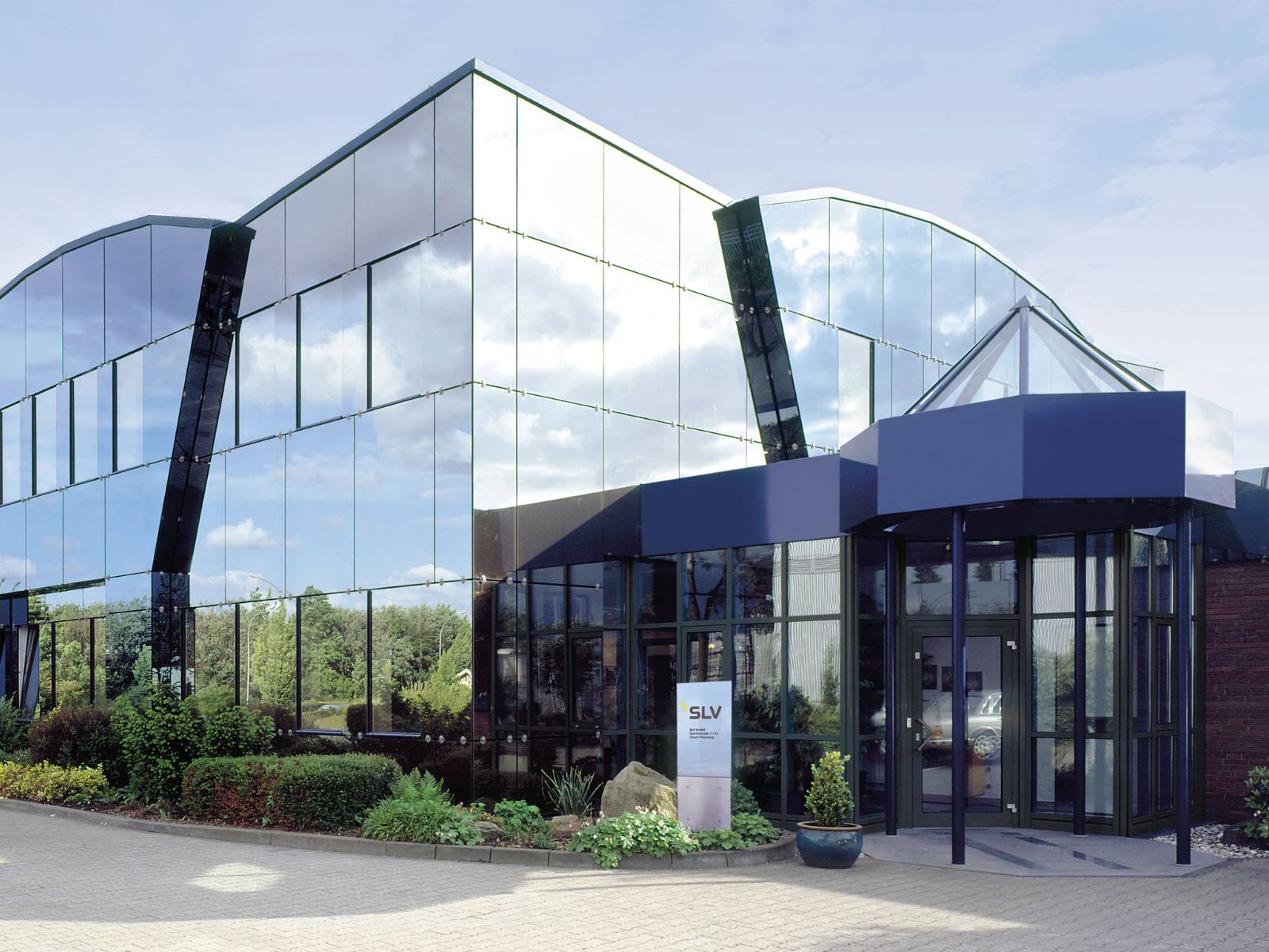 Der moderne Hauptsitz der SLV GmbH in Übach-Palenberg mit einer großen, spiegelnden Glasfassade, einem blauen Eingangsbereich und einer SLV-Firmensäule im Vordergrund.