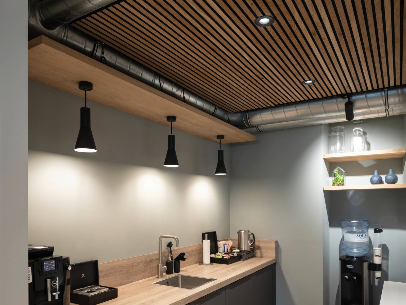 Schwarze runde Einbau-Downlights integriert in eine Holzlamellendecke über einer modernen Kaffeeküche mit zusätzlichen Pendelleuchten