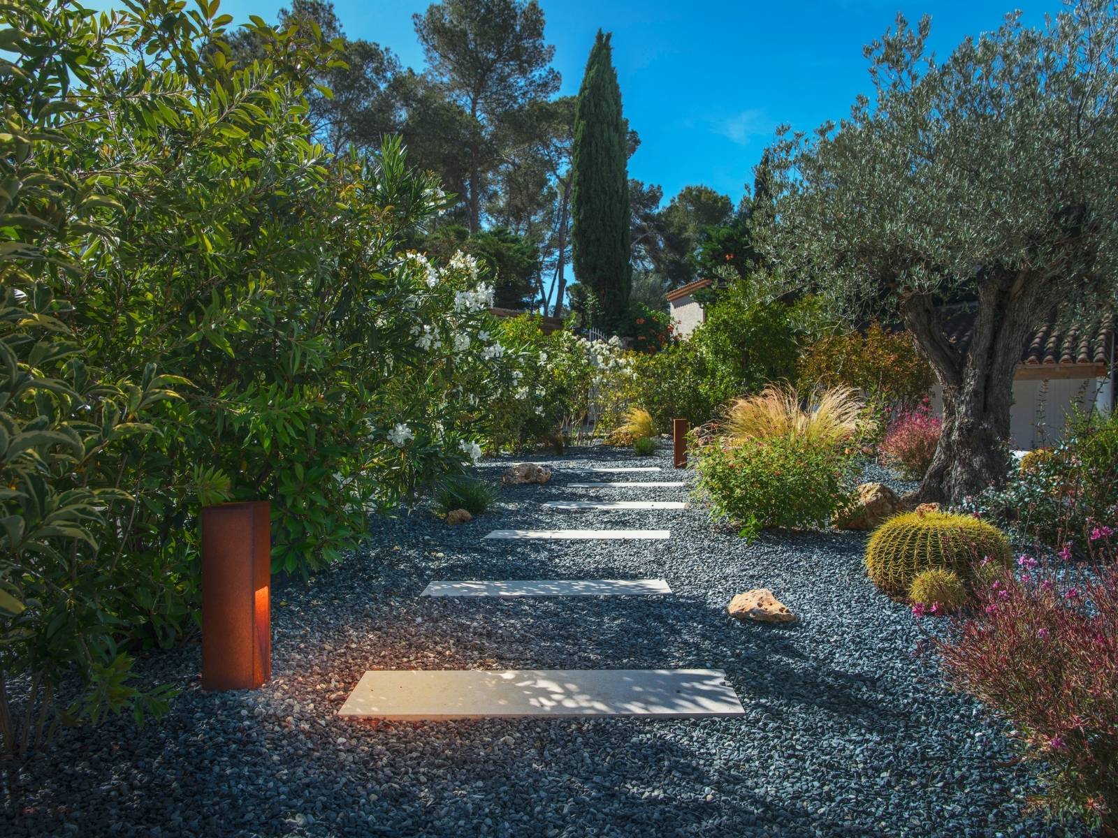 Mediterraner Garten mit einem Plattenweg im dunklen Kiesbett, umgeben von Kakteen und Sträuchern, beleuchtet durch rostfarbene LED-Pollerleuchten in Rost-Optik.