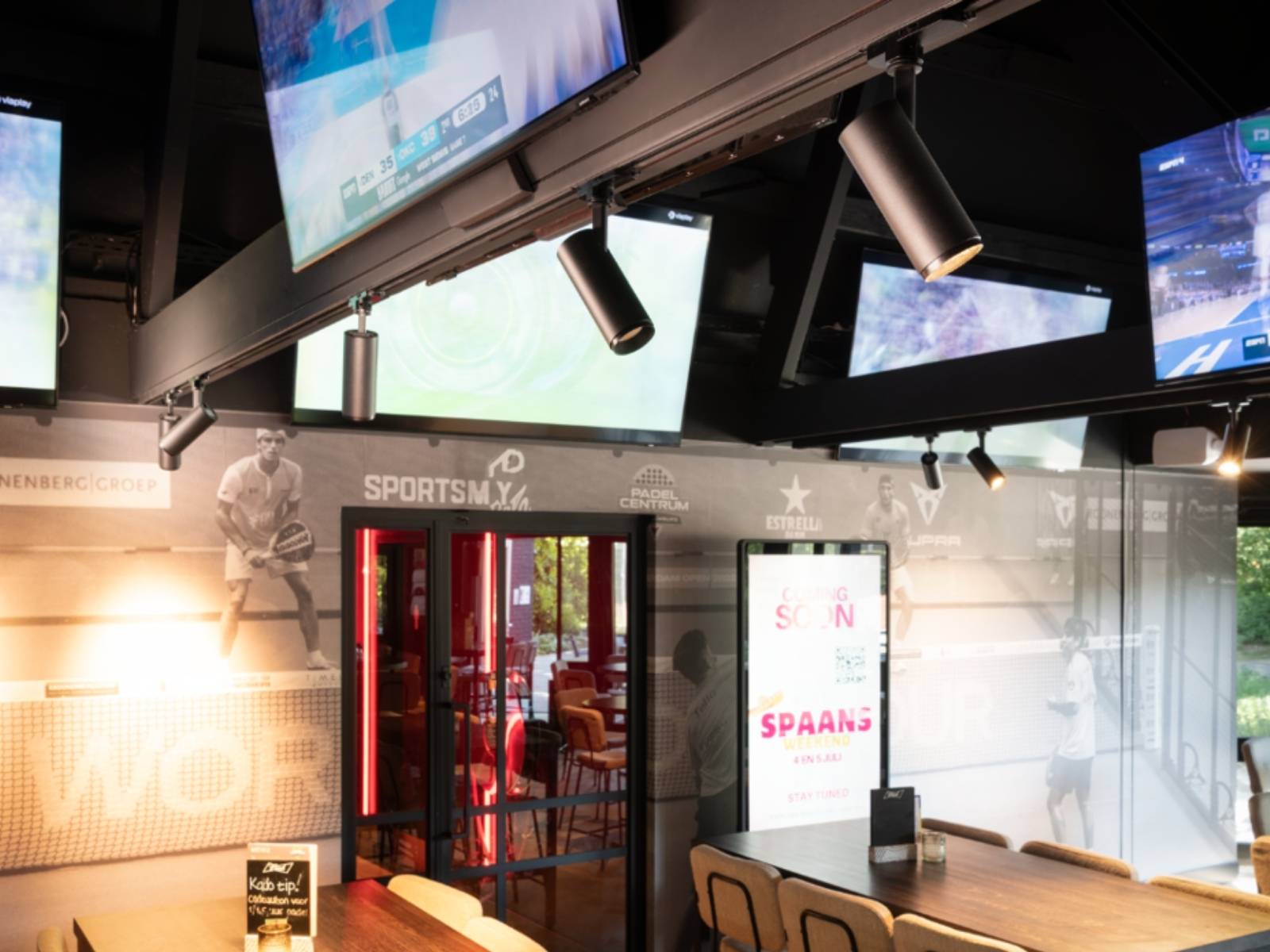 Innenausstattung einer Sportbar mit mehreren Bildschirmen, auf denen Spiele übertragen werden, Trophäenauslage, Holztischen und Stühlen. Hell beleuchtet durch NUMINOS® 3-Phasen-Spots mit Blick nach draußen durch Fenster.