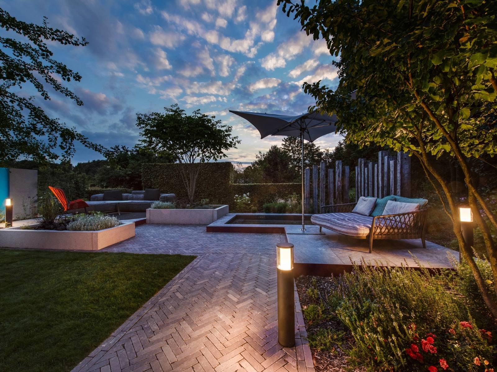 Modern gestaltete Hotelterrasse und Gartenanlage am Abend mit gemütlichen Loungemöbeln, Sonnenschirm und Pollerkleuchten, die den gepflasterten Weg sanft illuminieren.