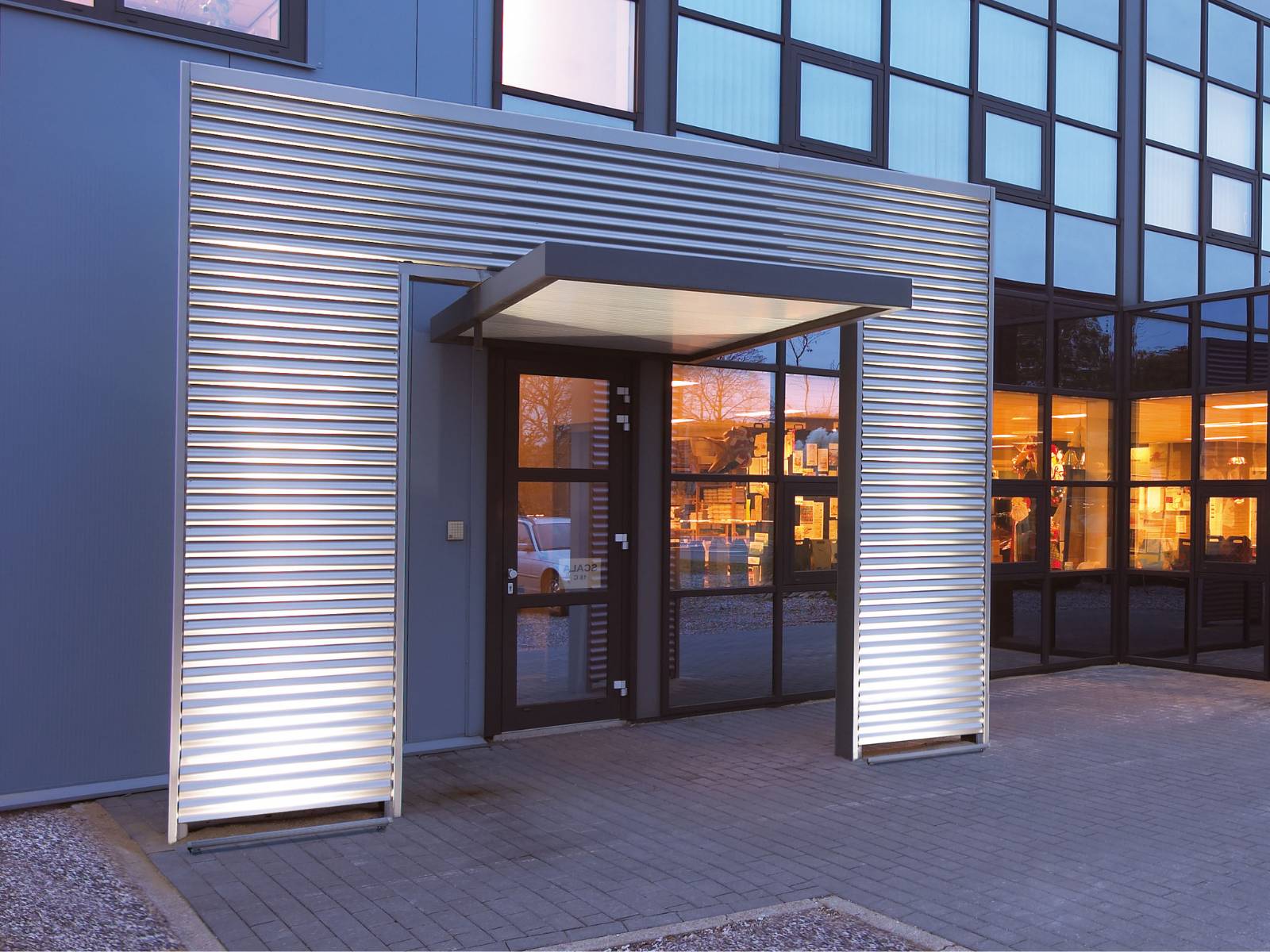 Eingangsbereich mit einer Fassade aus Lamellen, die durch integrierte Lichtpaneele und ein beleuchtetes Vordach in Szene gesetzt wird