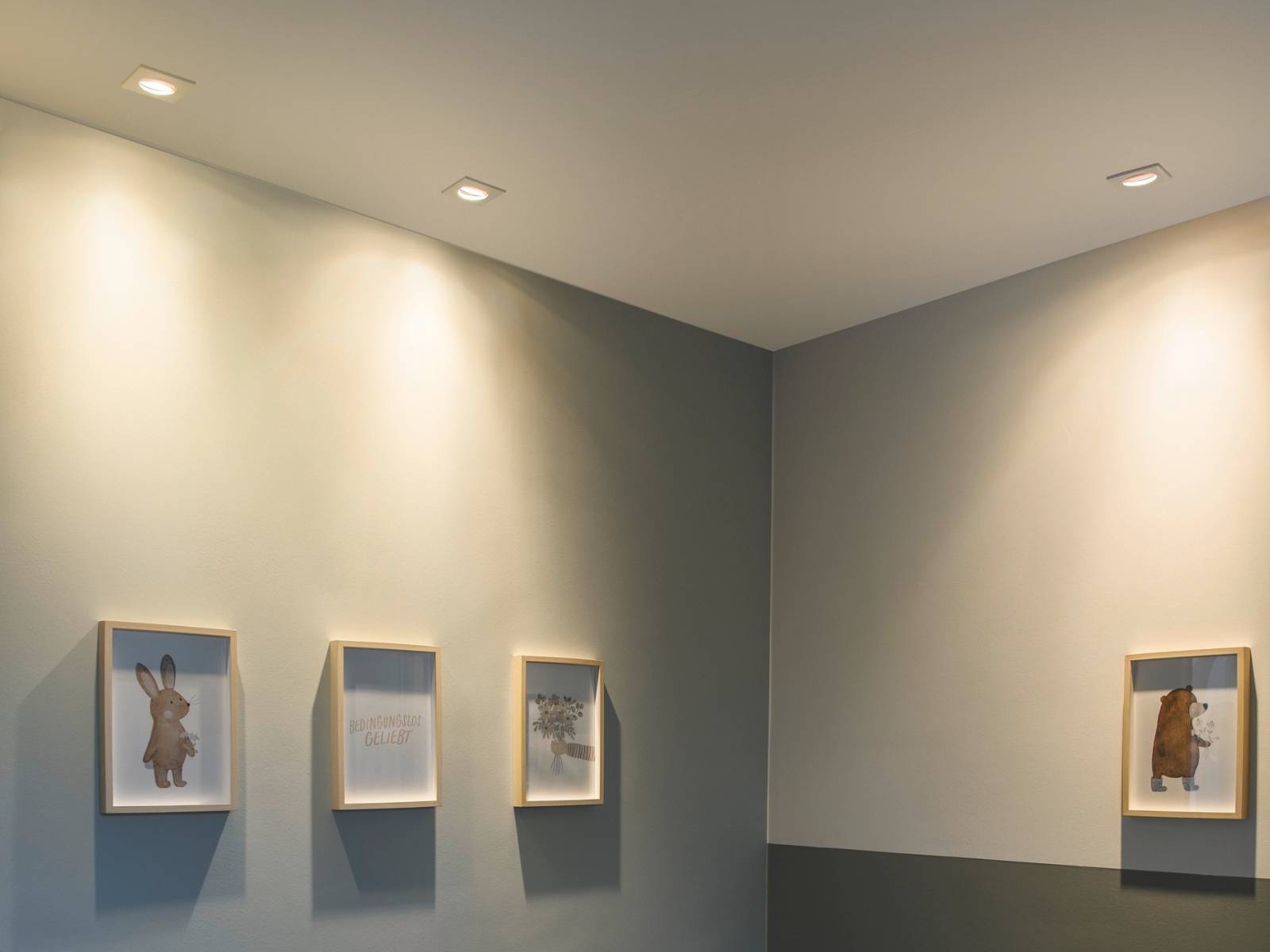 Quadratische weiße Downlights setzen als Akzentbeleuchtung kleine gerahmte Kunstwerke an einer hellgrauen Wand in Szene