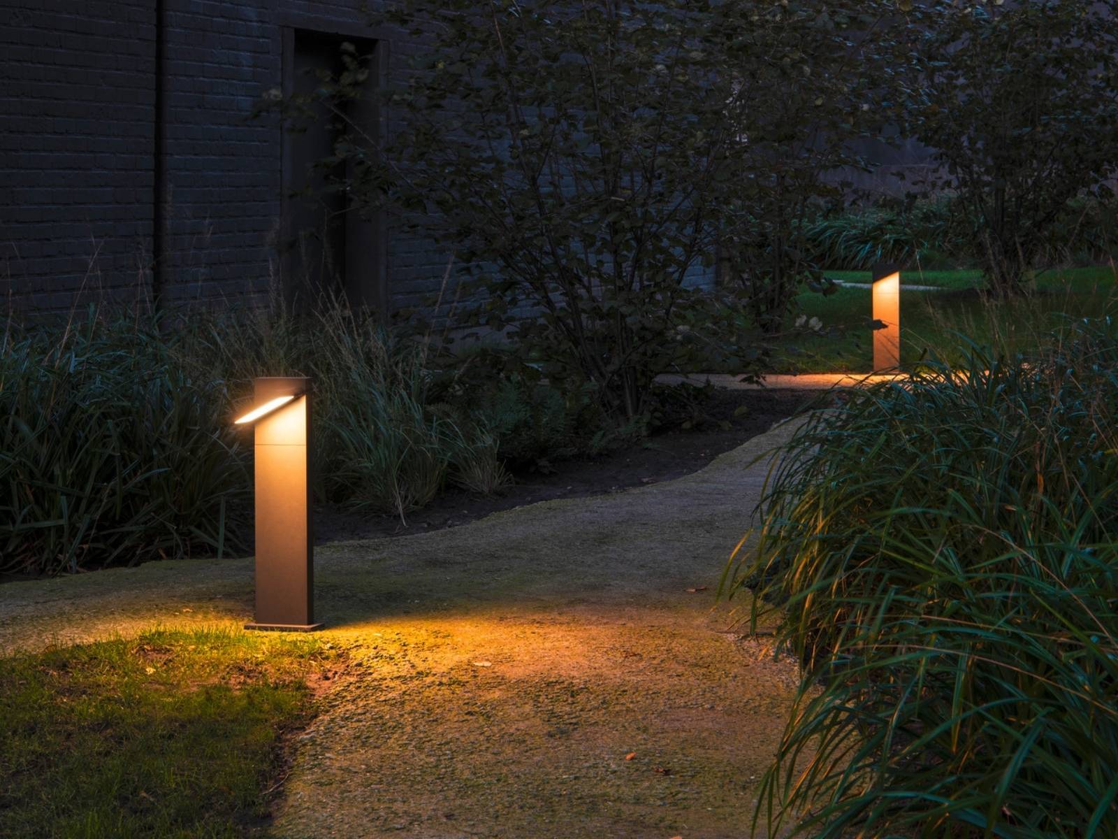 Zwei moderne, quaderförmige Wegeleuchten aus dunklem Metall beleuchten mit warmem LED-Licht einen geschwungenen Gartenweg in der Abenddämmerung.