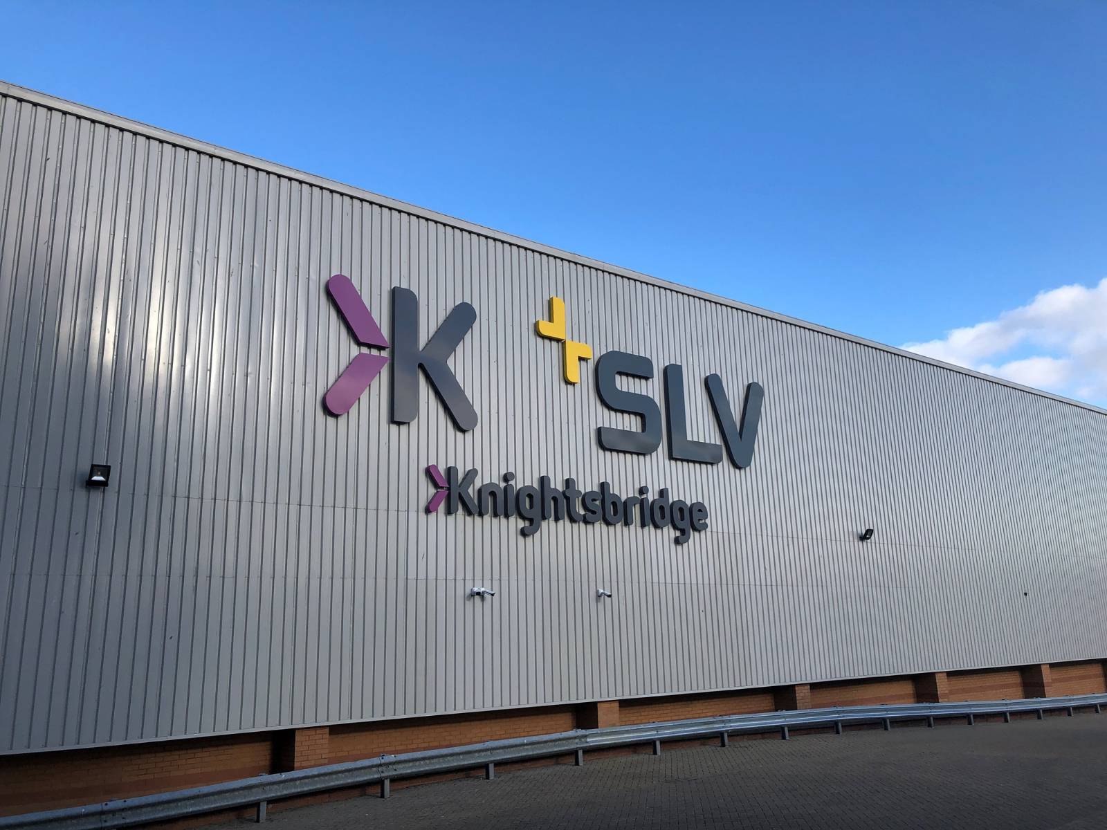 The metallic grey exterior façade of the UK site, featuring the Knightsbridge and SLV logos mounted side by side, against a blue sky.