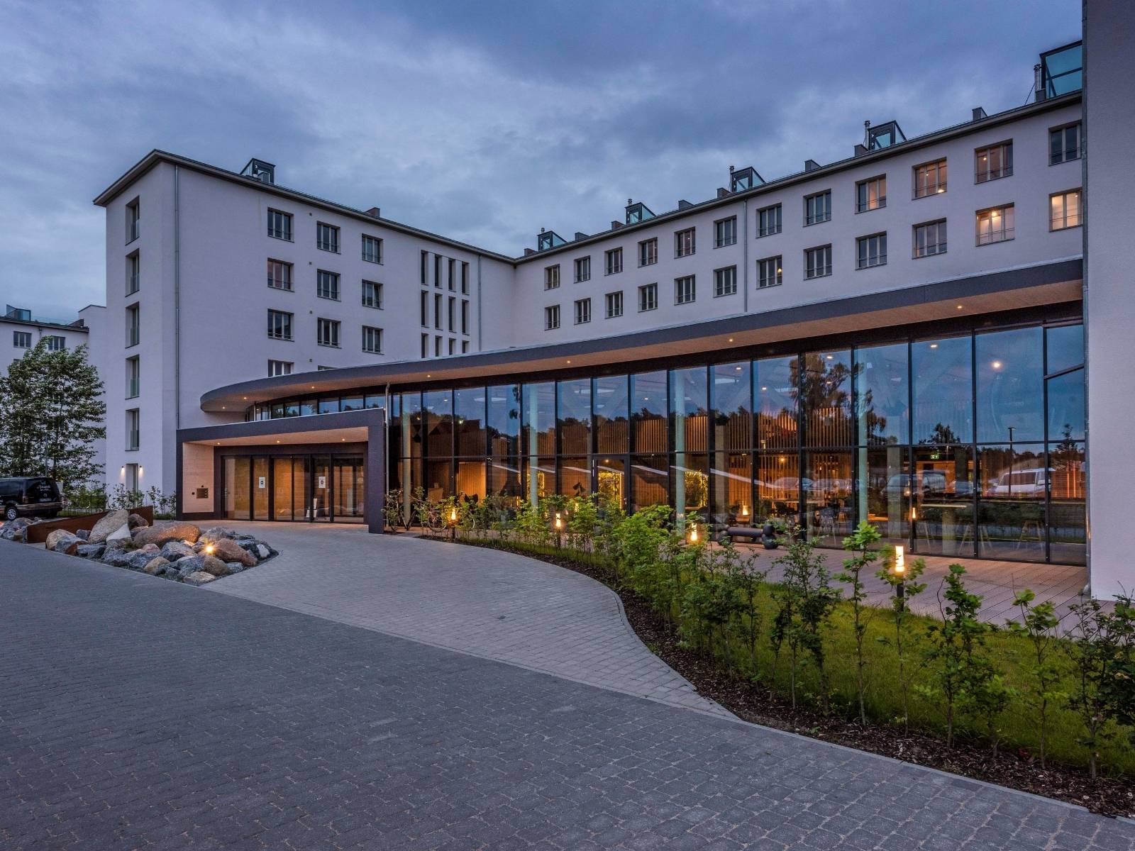 Großzügiger Eingangsbereich eines modernen Hotels in der Dämmerung mit einer langen Glasfront, gepflasterter Vorfahrt und dezenten Pollerleuchten im Grünstreifen.