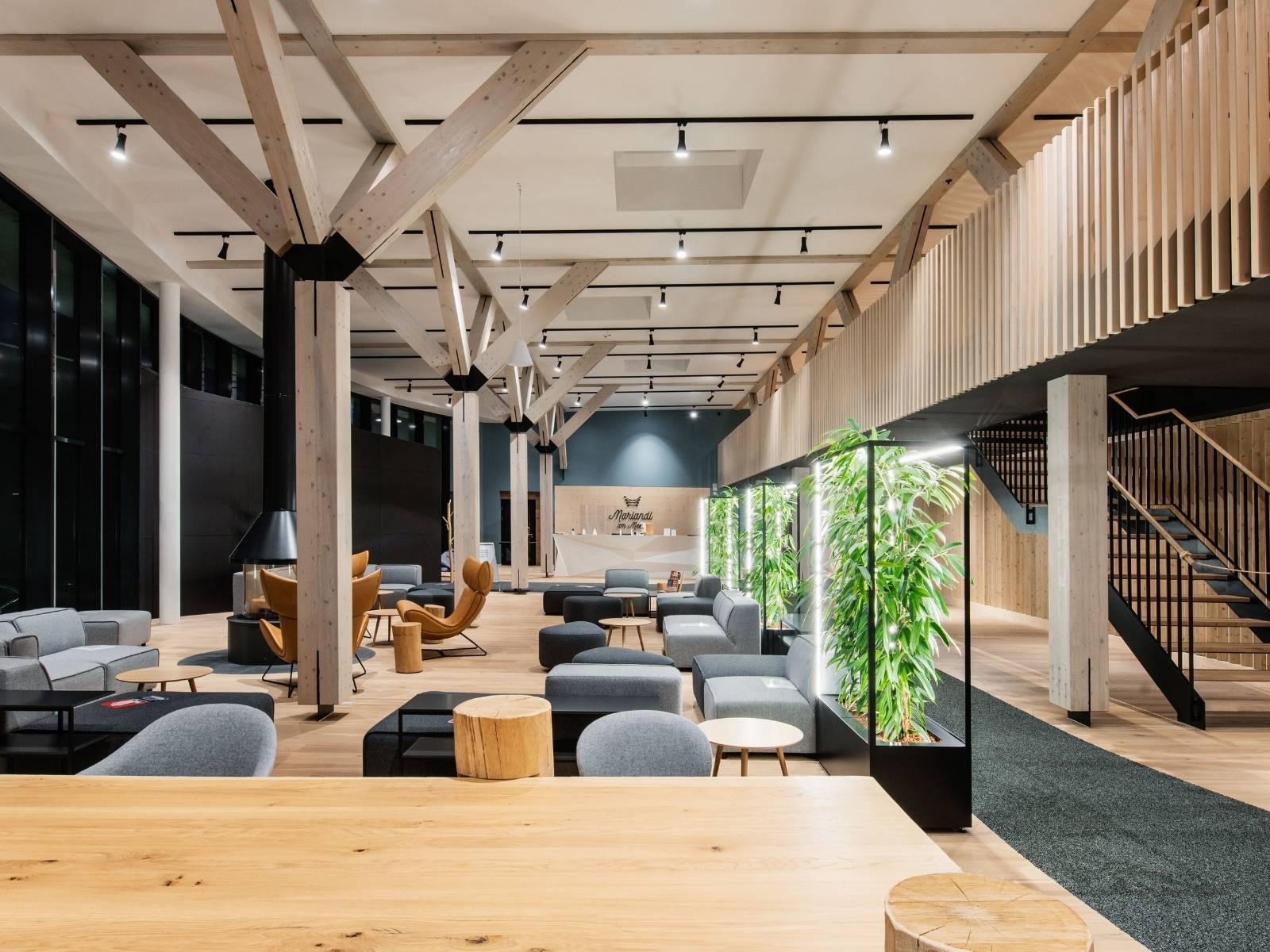 Großzügige, offen gestaltete Hotel-Lobby im alpinen Stil mit hellen Holzdeckenbalken, grauen Polstersesseln, einem Kamin und Pflanzenelementen mit integrierter LED-Beleuchtung.