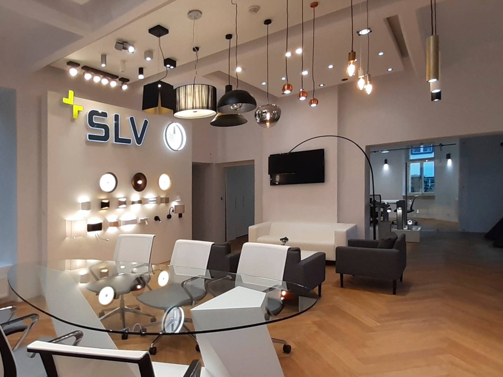 Moderní showroom svítidel se skleněným konferenčním stolem v popředí, osvětleným logem SLV na stěně, ukázkami nástěnných lamp a řadou různých designových závěsných lustrů.