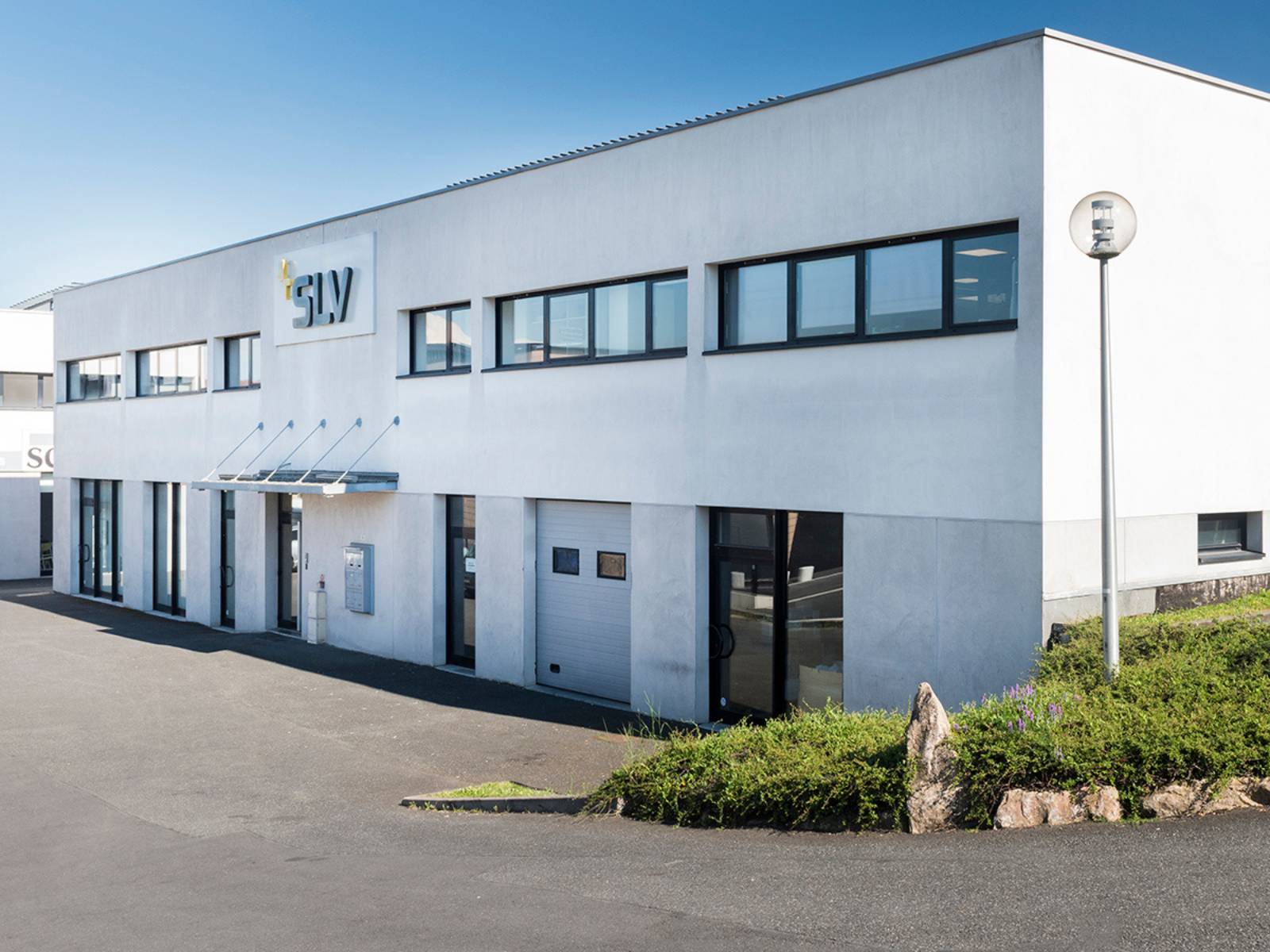 Façade moderne et blanche du bâtiment de SLV France par une journée ensoleillée, avec le logo de l'entreprise au-dessus de l'entrée principale, de larges fenêtres et une entrée de garage intégrée.