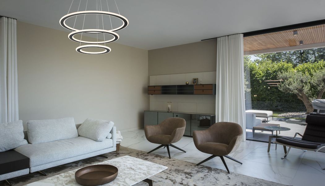 Modernes Wohnzimmer mit ONE TRIPLE Pendelleuchte, weißem Sofa, braunen Stühlen, Marmortisch und großen Fenstern, die sich zum Garten hin öffnen.