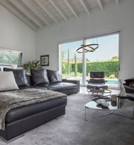 Modernes Wohnzimmer mit schwarzem Ecksofa, Glas-Couchtisch mit ONE DOUBLE Pendelleuchte und großen Schiebetüren, die den Blick auf den Pool und den Garten freigeben.