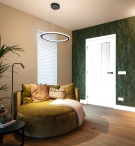 Gemütliches Zimmer mit einer grünen Akzentwand, einem runden grünen Sofa, Pflanzen, einem Beistelltisch und ONE FLAT Pendelleuchte. Heller Holzboden und weiße Tür.