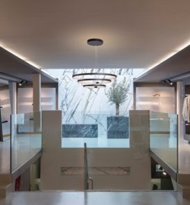Modernes Interieur mit einer zentralen Treppe, Glasgeländern, ONE TRIPLE Pendelleuchte und einem großen Fenster, durch das man einen Baum draußen sehen kann.