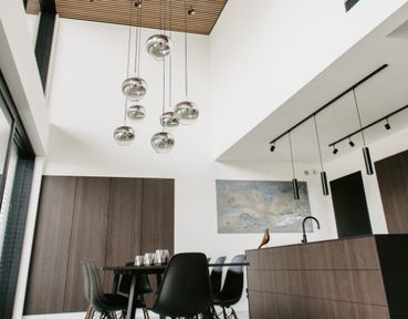 Moderne Küche und Essbereich mit Holzmöbeln, eleganten schwarzen Arbeitsplatten, Pendelleuchten in Metallic-Optik und einer hohen Decke.