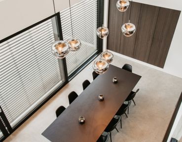 Moderner Essbereich mit einem langen schwarzen Tisch, schwarzen Stühlen und hängenden kugelförmigen Pendelleuchten. Große Fenster mit Jalousien lassen den Raum hell erscheinen.