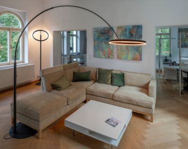 Modernes Wohnzimmer mit einem beigen Ecksofa, grünen Kissen, einem weißen Couchtisch und der hohen, geschwungenen ONE BOW Stehleuchte. Große Fenster und abstrakte Kunst.