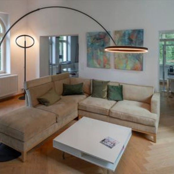Modernes Wohnzimmer mit einem beigen Ecksofa, grünen Kissen, einem weißen Couchtisch und der hohen, geschwungenen ONE BOW Stehleuchte. Große Fenster und abstrakte Kunst.
