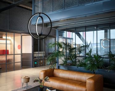 Moderne Büro-Lobby mit einem braunen Ledersofa, Glaswänden, Zimmerpflanzen und der ONE VERTICAL PD, die von der Decke hängt.