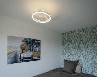 Modernes Schlafzimmer mit der ONE Wand- und Deckenaufbauleuchte, einer Tapete mit Blattmuster, einem Bett mit braunen Kissen und einem Gemälde mit einem Oldtimer an der Wand.