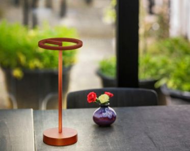 Die VINOLINA ONE Akku-Leuchte auf einem Tisch mit einer kleinen violetten Vase mit roten und rosa Blumen vor einem verschwommenen Hintergrund im Freien.