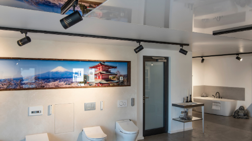 Moderner Badezimmer-Showroom mit Toiletten, Waschbecken und einem Panoramafoto einer japanischen Landschaft an der Wand. Der Raum wird durch Schienenbeleuchtung erhellt.