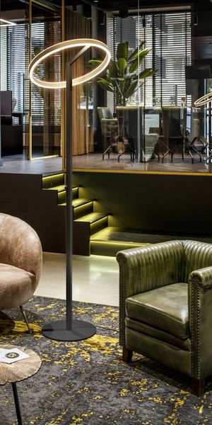 Modernes Interieur mit der ONE-Stehleuchte, Ledersesseln, einem gemusterten Teppich und einem Konferenzraum mit Glaswänden im Hintergrund.