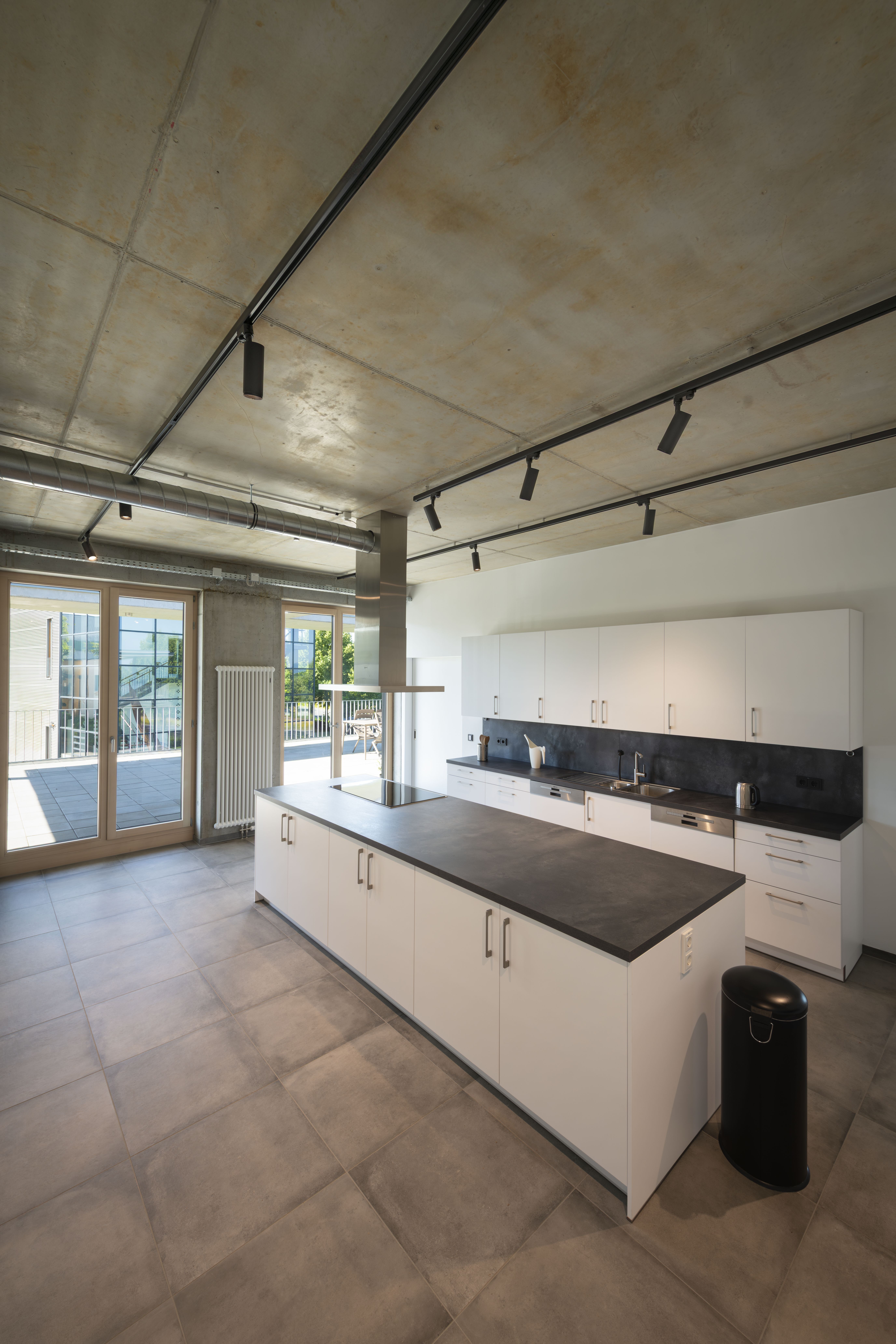 Moderne Küche mit weißen Schränken, schwarzen Arbeitsplatten und einer großen Kochinsel. Sichtbetondecke und S-TRACK Schienen mit NUMINOS® Beleuchtung. Große Fenster auf der linken Seite.