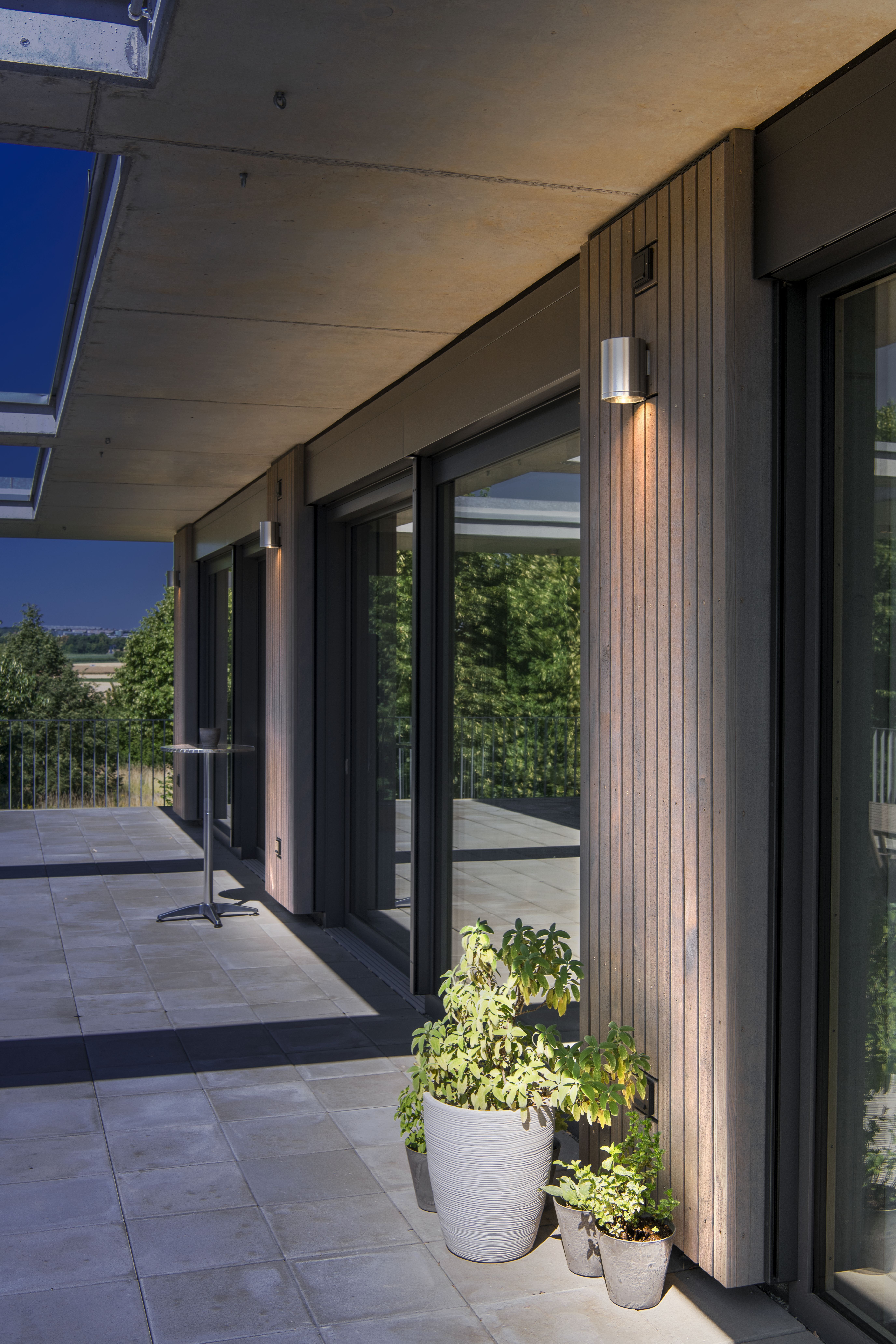 Moderne Terrasse mit großen Glastüren, Topfpflanzen, einer Holzfassade und ROX UP/DOWN Wandleuchte. Ein malerischer Blick auf Bäume im Hintergrund.