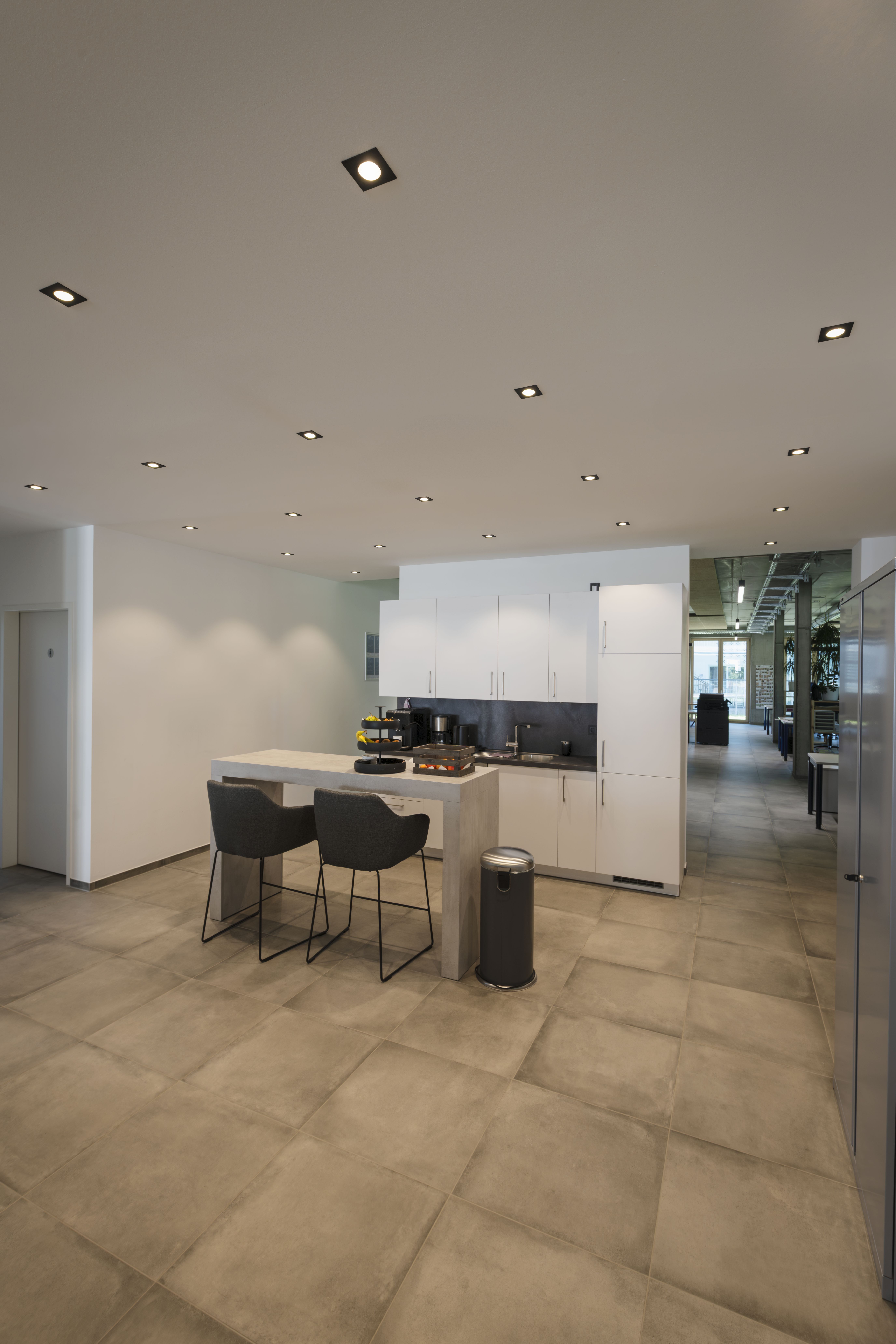 Moderne Küche mit weißen Schränken, einer zentralen Kochinsel mit zwei grauen Stühlen und NEW TRIA® Einbauleuchten. Grauer Fliesenboden und elegante Küchengeräte.