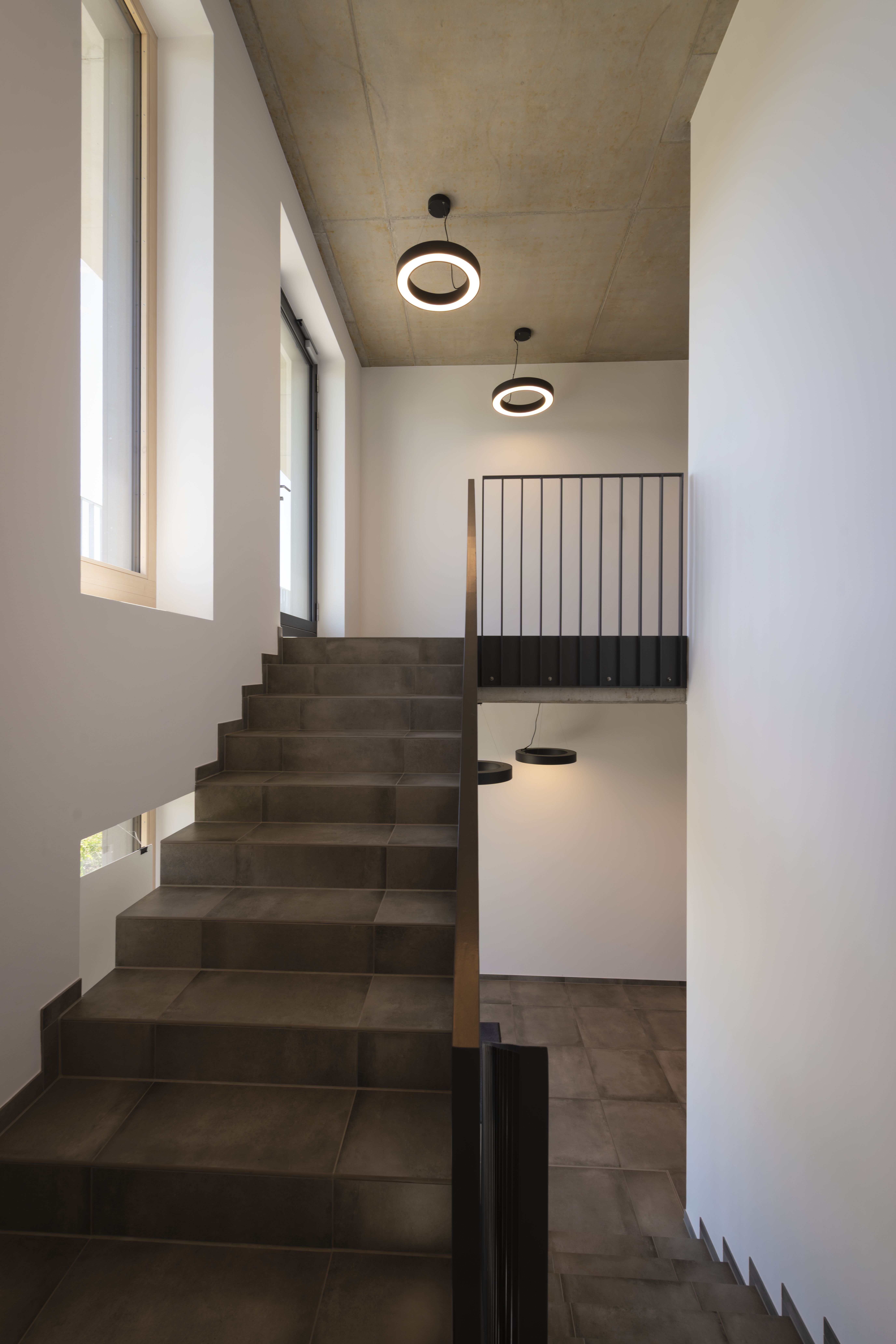 Modernes Interieur mit einer Treppe, dunkel gefliesten Stufen, schwarzen Geländern, großen Fenstern und MEDO® RING Pendelleuchten.