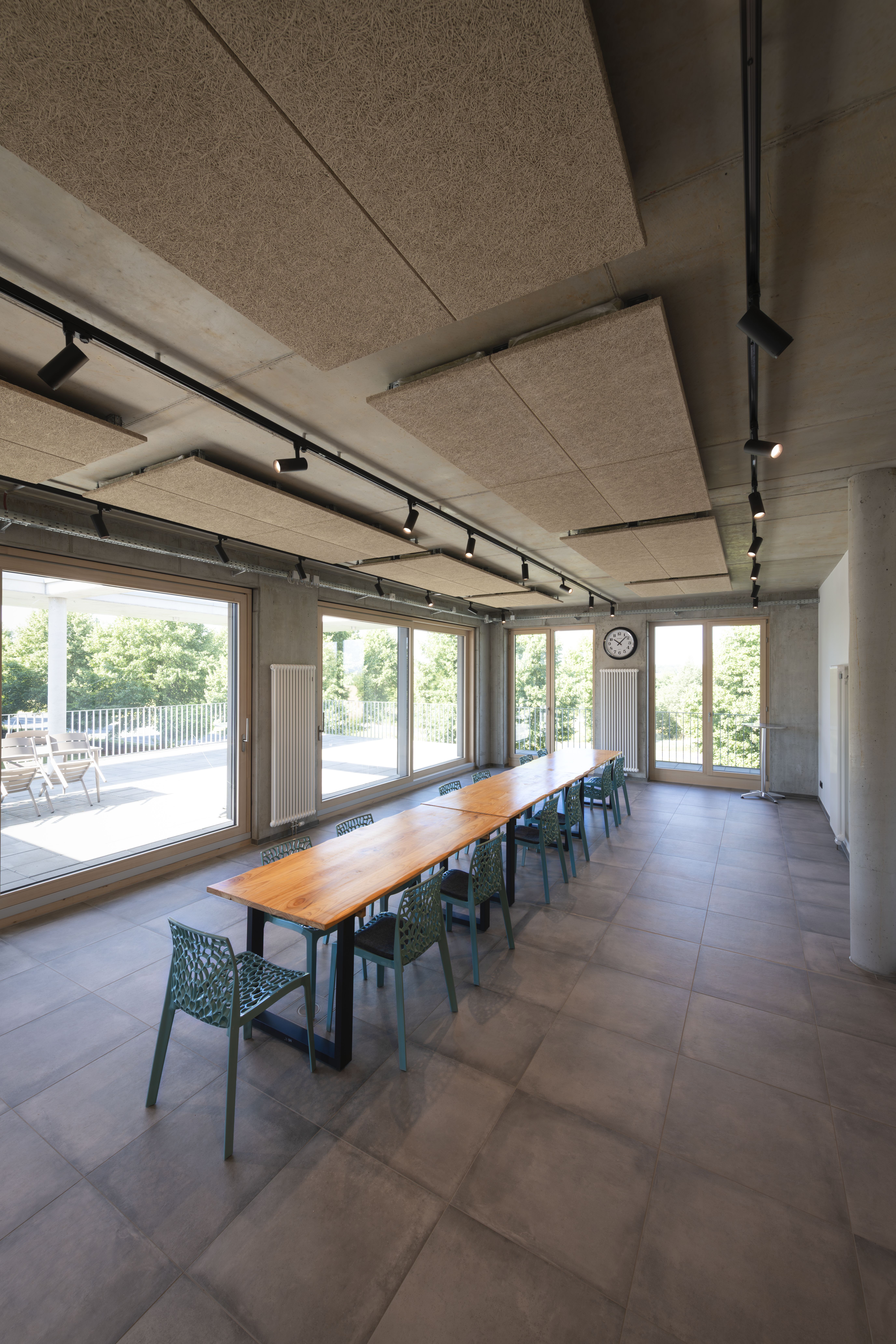 Ein moderner Konferenzraum mit einem langen Holztisch - beleuchtet von NUMINOS® Deckenspots- , grünen Stühlen, großen Fenstern und einer Wanduhr, mit Blick auf einen üppig begrünten Außenbereich.